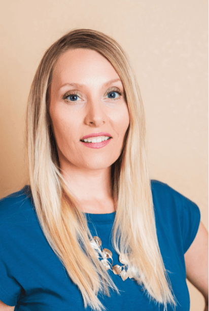 Smiling blonde woman with blue eyes wearing a blue shirt and a gold necklace.
