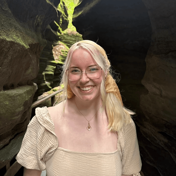 Smiling blonde woman with glasses and headscarf stands in a narrow, dark, mossy rock canyon.